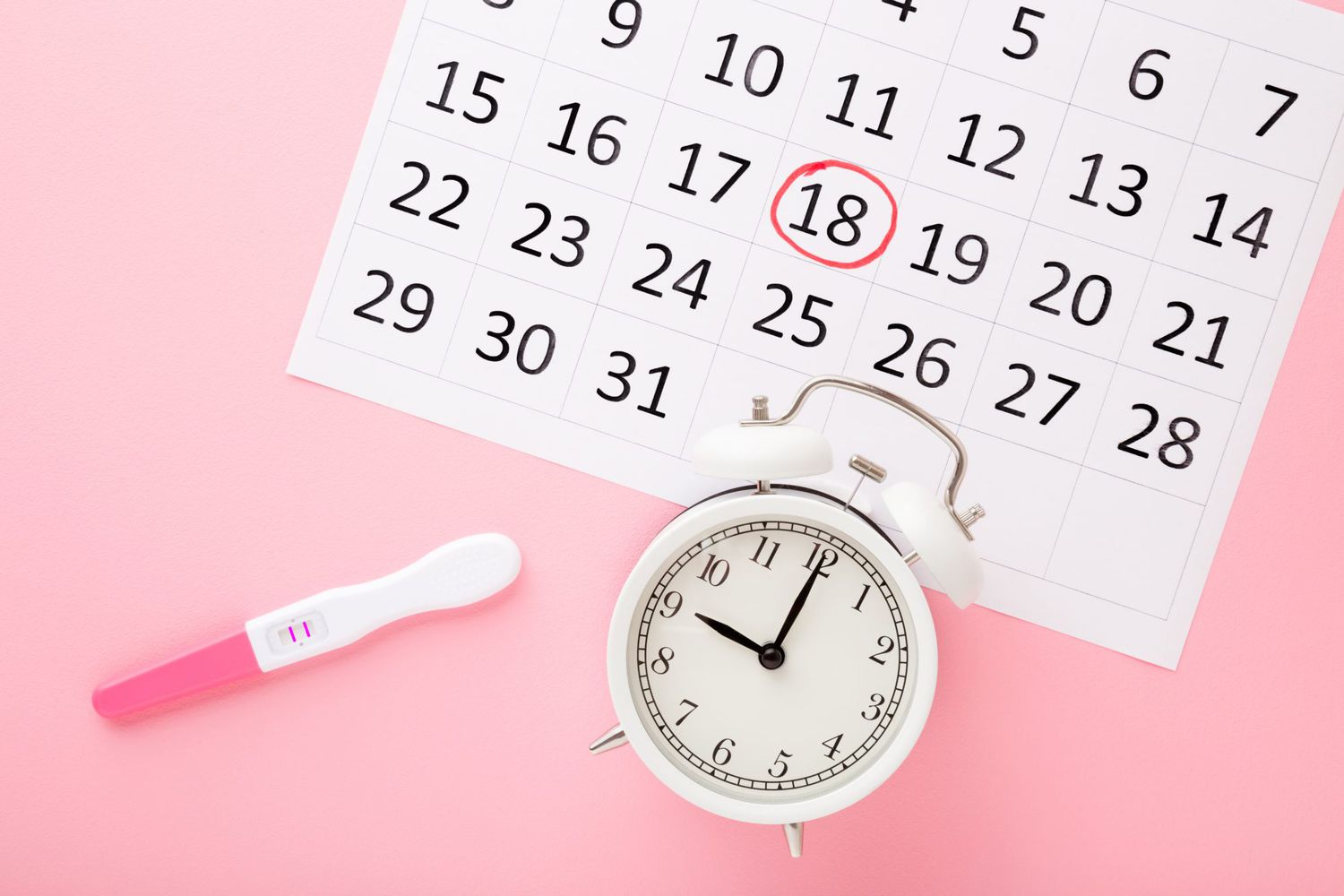 An image of a pregnancy test, calendar, and alarm clock on a pastel pink background.