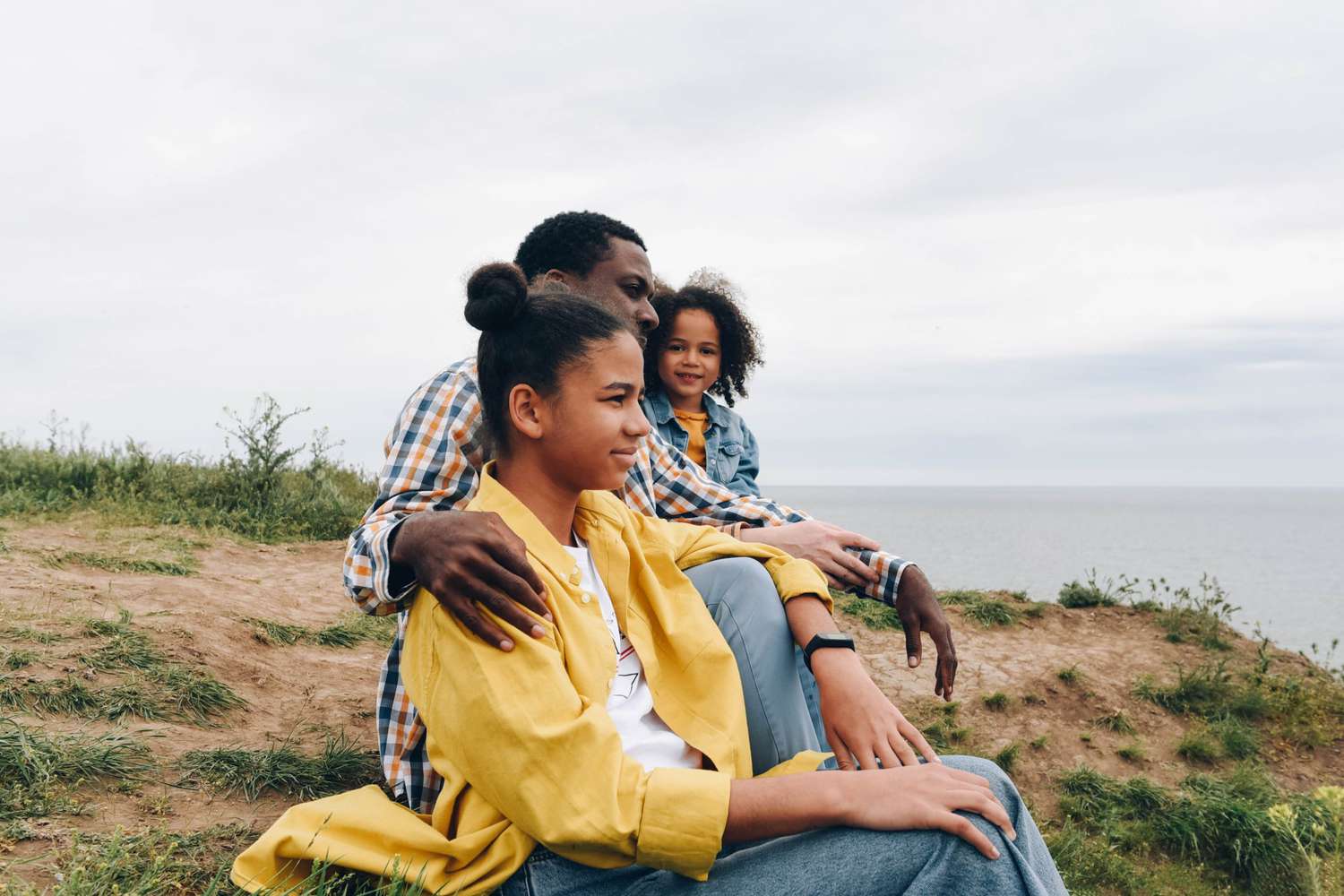Man and two daughters sit on hillside over looking the sea.
