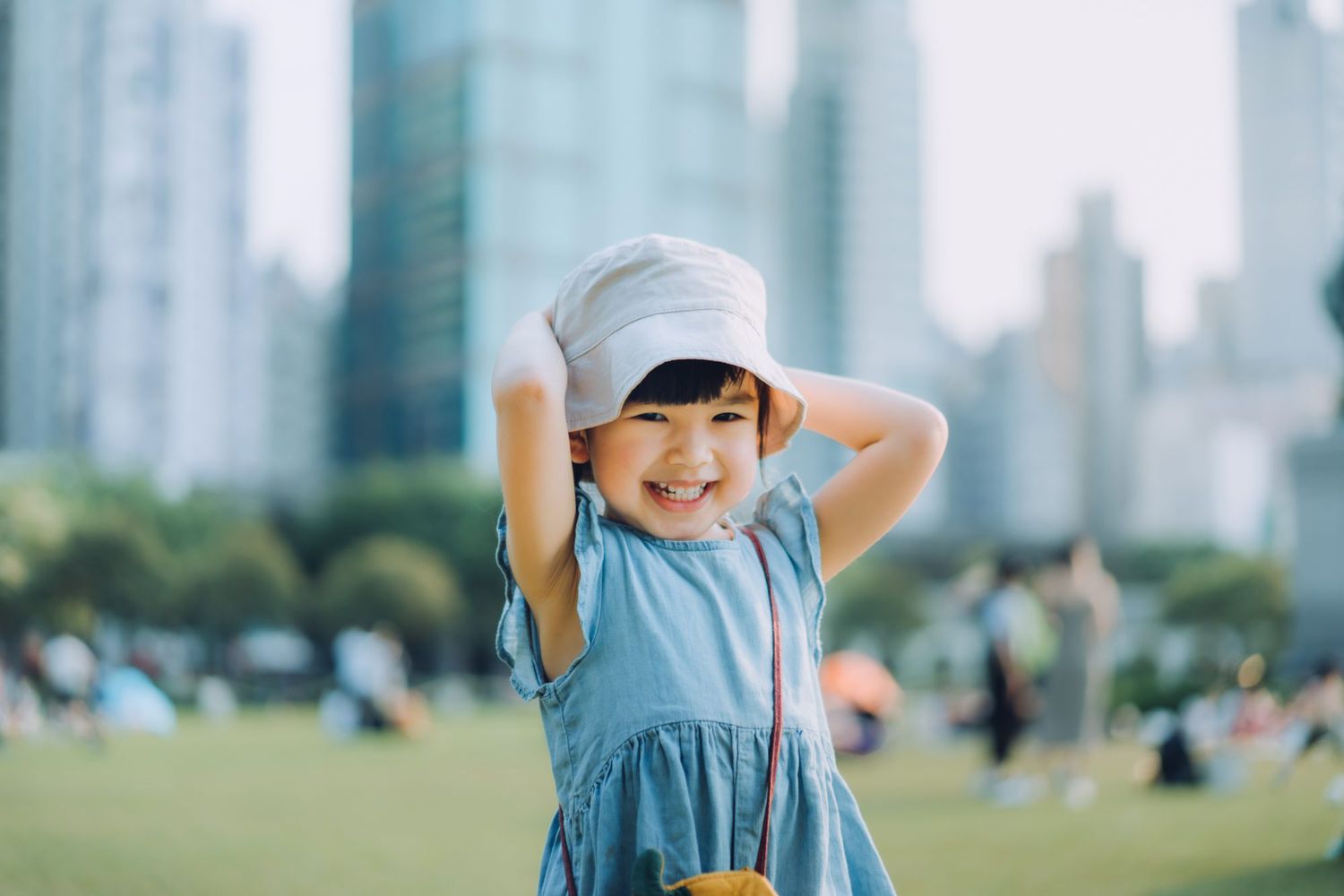 Portrait of happy little Asian girl having fun playing in urban park, smiling joyfully. Enjoying freedom and beauty of nature. With modern cityscape in background