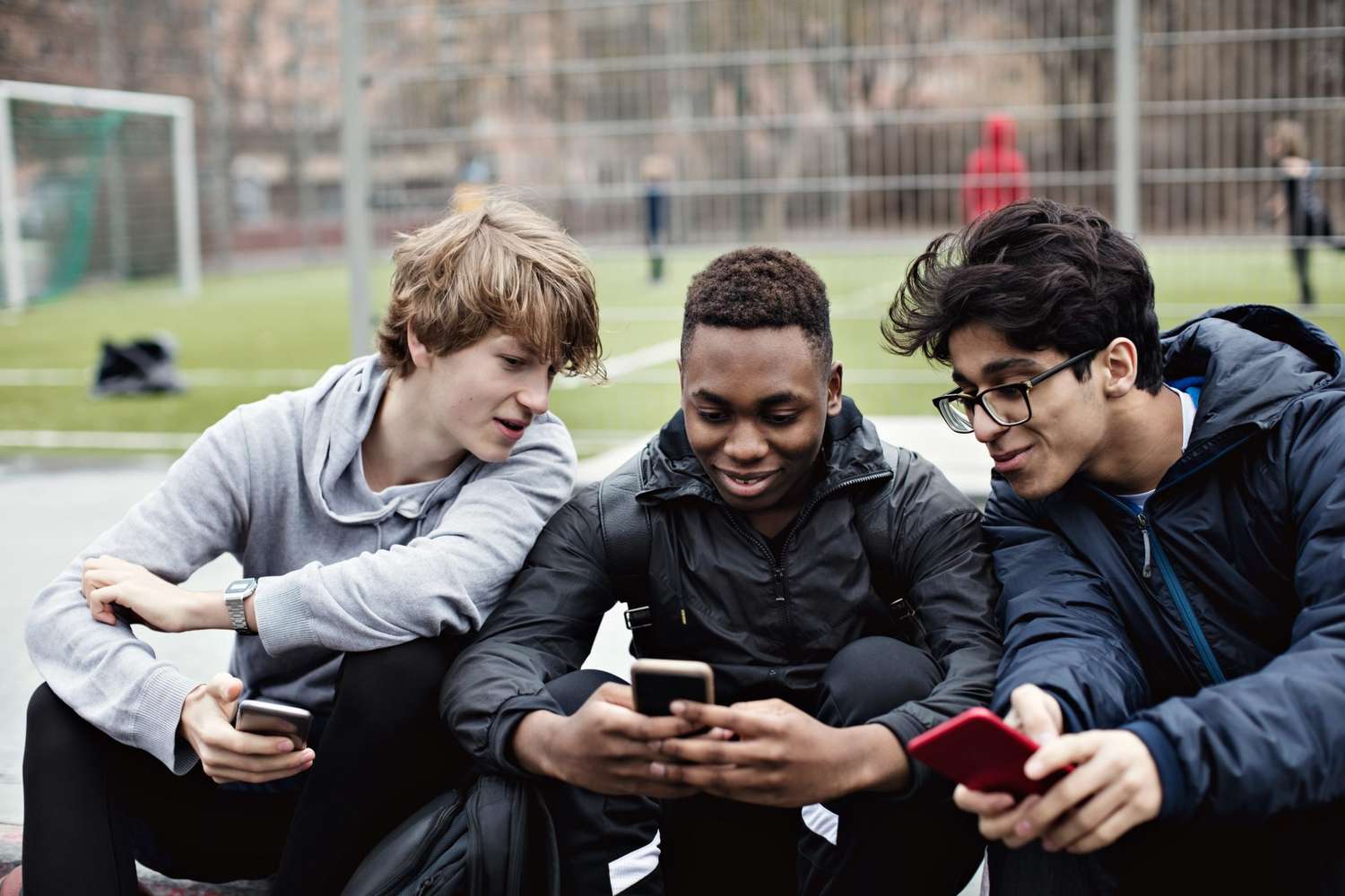 Smiling friends looking at teenage boy's phone while sitting against soccer field