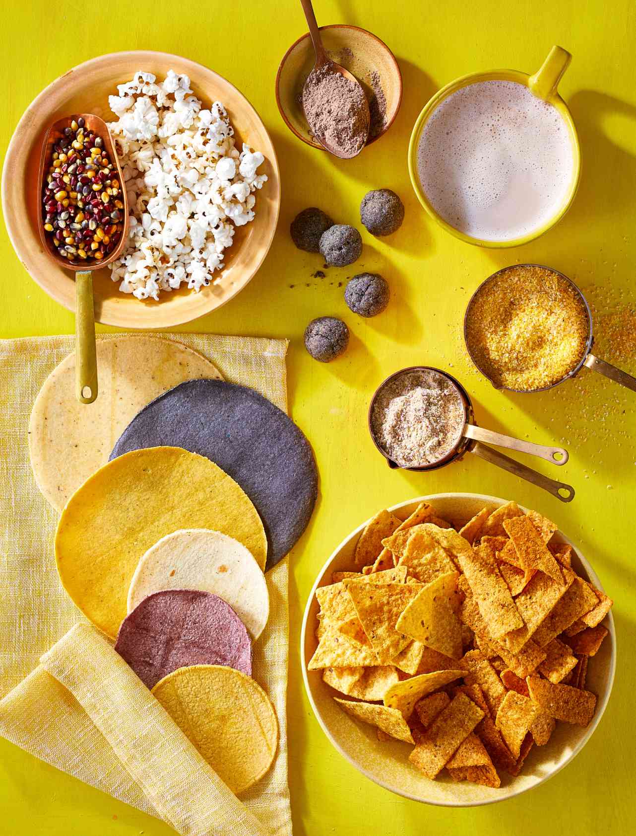 Assortment of corn products like flour, tortillas, chips and popcorn in bowls on yellow background.