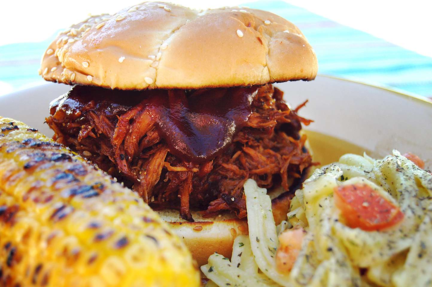 side view of a pulled pork sandwich with barbecue sauce on a plate with grilled corn on the cob and coleslaw
