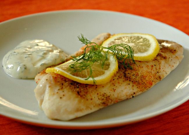 a baked tilapia fillet topped with lemon slices and a fresh dill sprig on a plate with tartar sauce