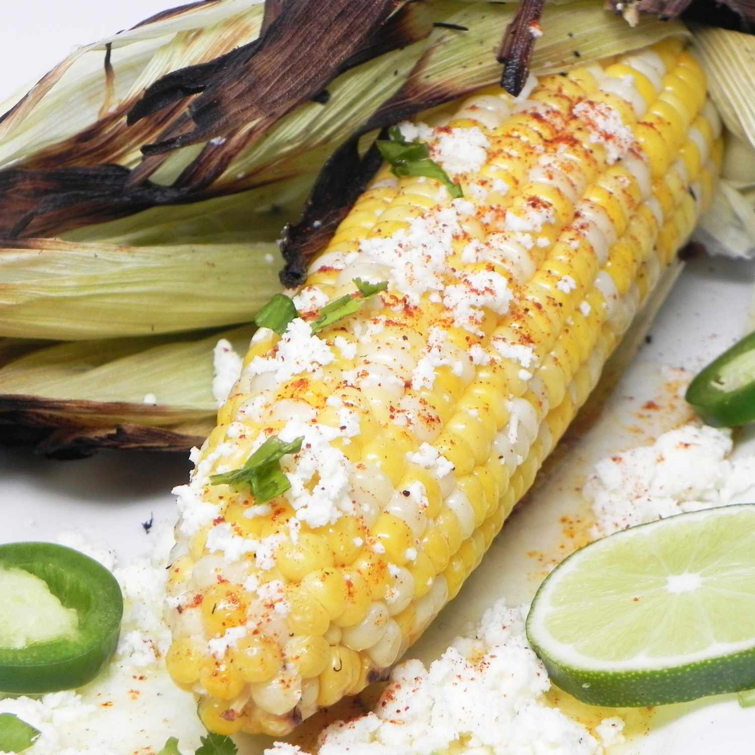closeup of cotija- and ground chile-topped corn on the cob with lime slices and charred husk on the side