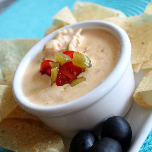 a small white bowl filled with queso, topped with chopped green and red chilies, with tortilla chips in the background