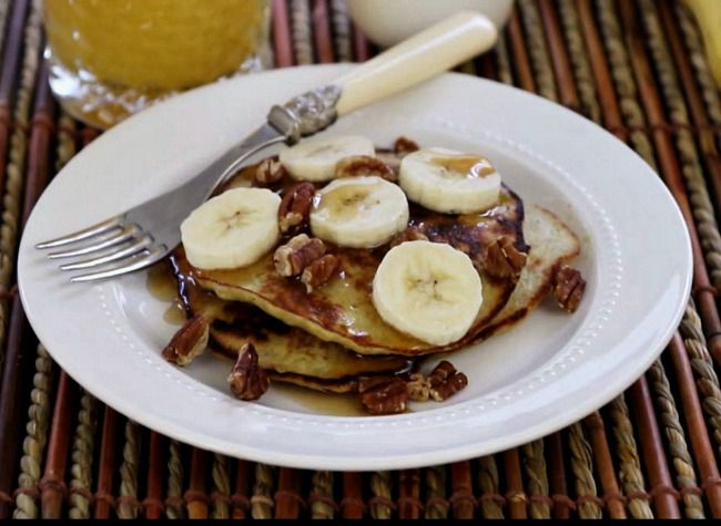 a white plate with a stack of pancakes topped with fresh banana slices and chopped pecans