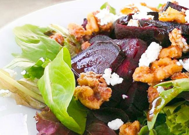 A closeup of sliced red beets, candied walnut pieces, and crumbled goat cheese on a bed of mesclun mix
