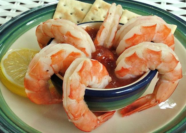 five shrimp arranged on a ramekin of cocktail sauce with a lemon wedge and crackers in the background