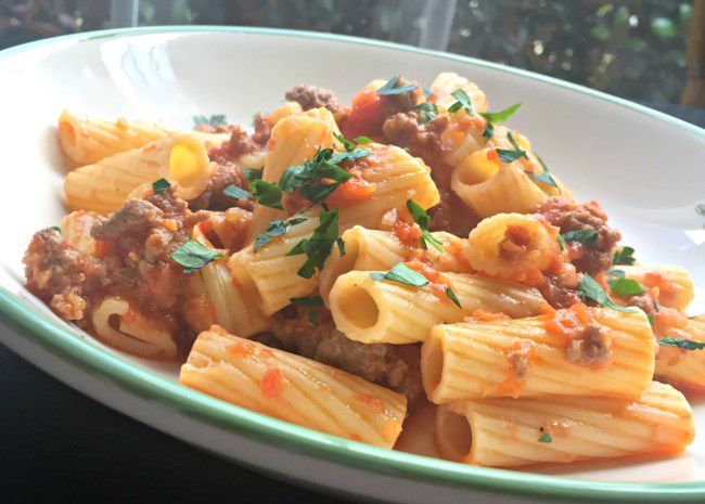 a bowl of rigatoni rigate topped with Bolognese sauce, garnished with chopped parsley