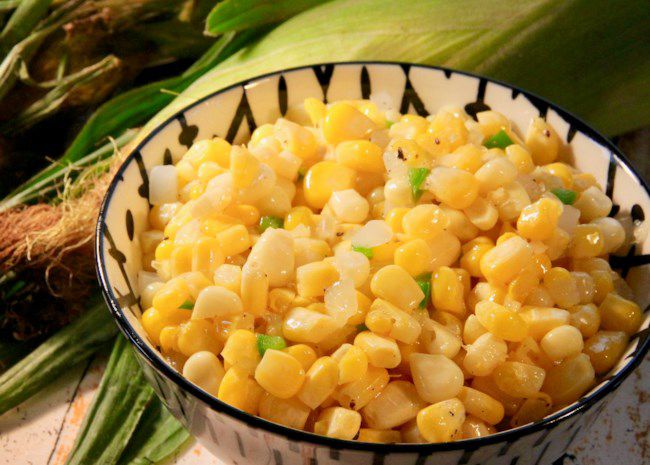 a small bowl of corn with corn on the cob in its husk in the background
