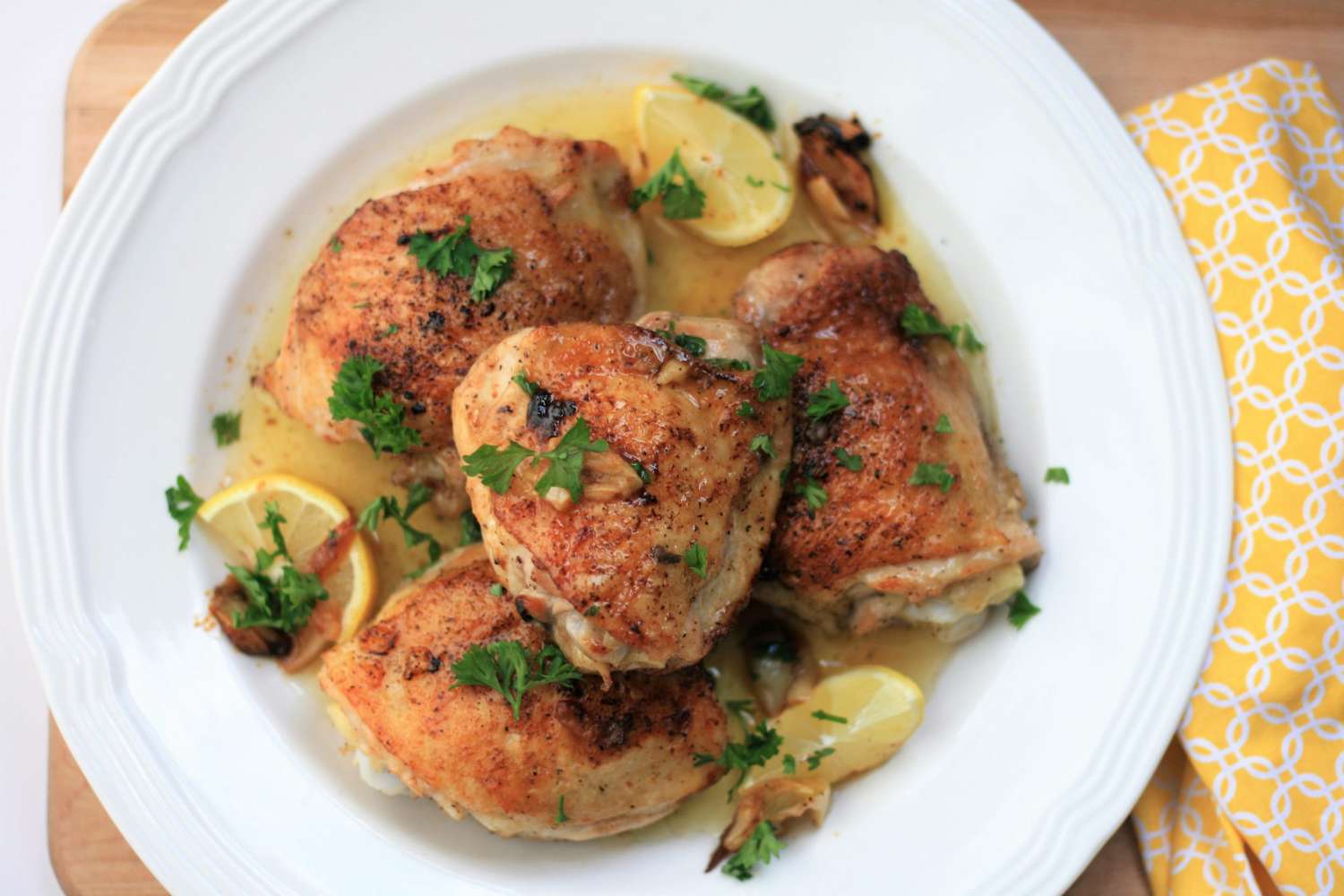 a top-down view of golden brown chicken thighs in a buttery sauce, garnished with lemon slices and chopped parsley