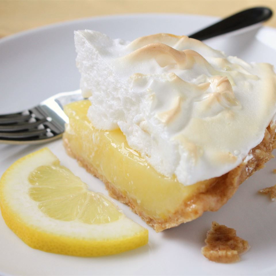 a slice of pie with lightly browned meringue, garnished with half a lemon slice on a white plate