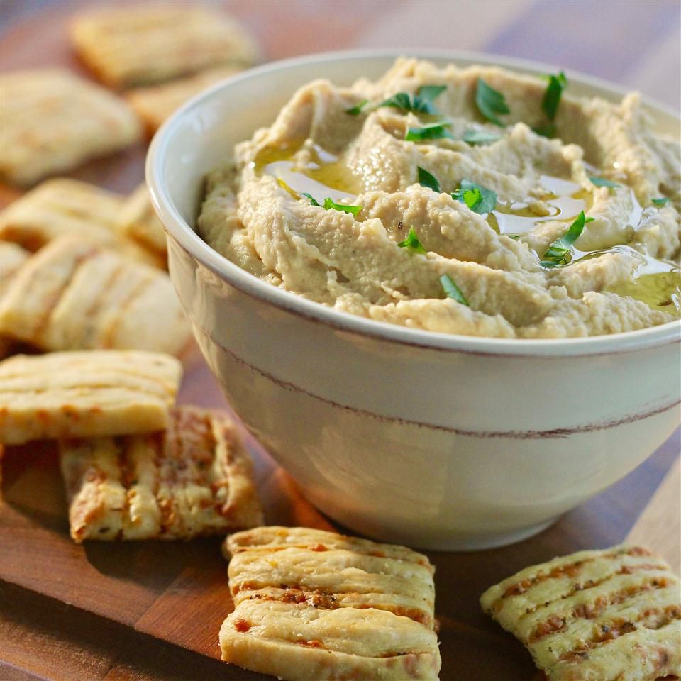 A small white bowl of parsley-topped hummus surrounded by pita squares