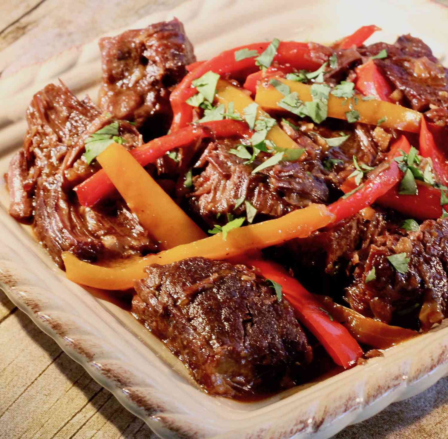 a square ceramic platter holding pieces of beef pot roast with slices of red and yellow bell pepper