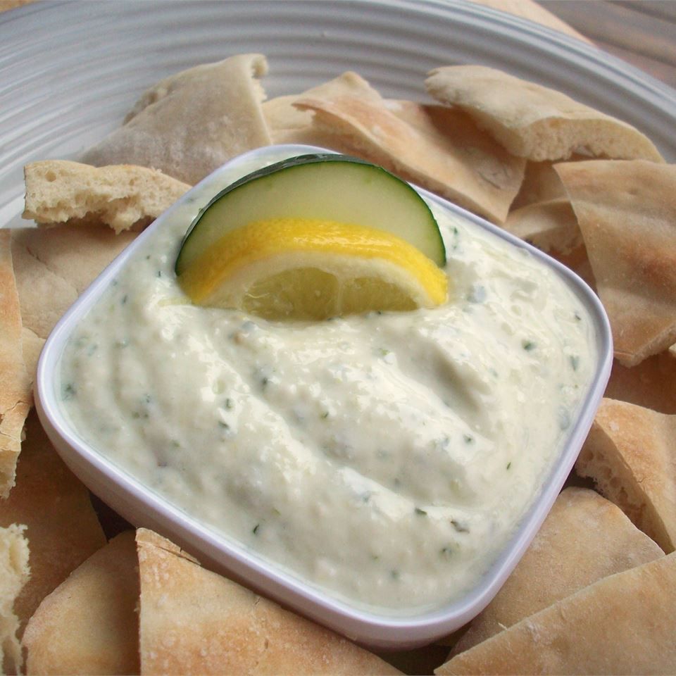 Cream white dip in a bowl with a lemon and cucumber slice. Around the bowl is pita wedges