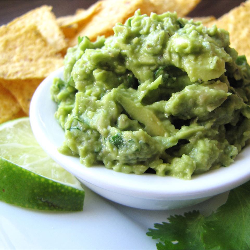 a small white bowl piled high with coarsely mashed avocado, with a lime wedge on the side and tortilla chips in the background