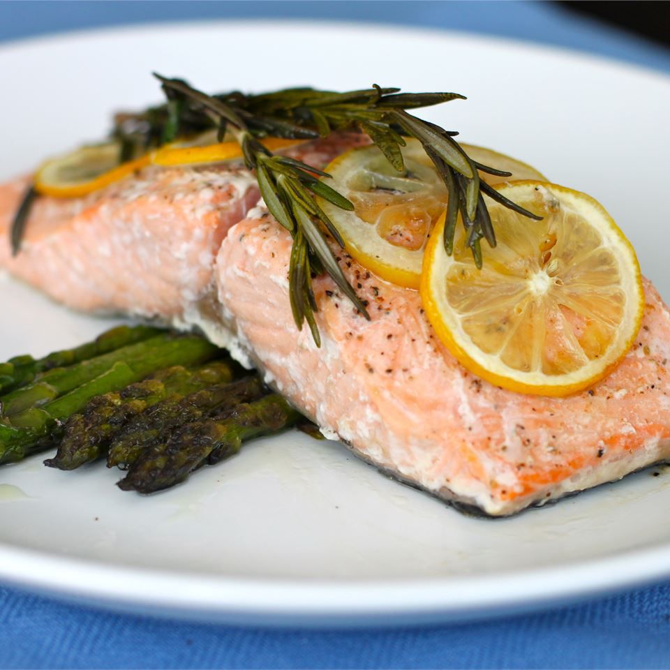 a plate with a baked salmon fillet on top of roasted asparagus, garnished with lemon slices and rosemary