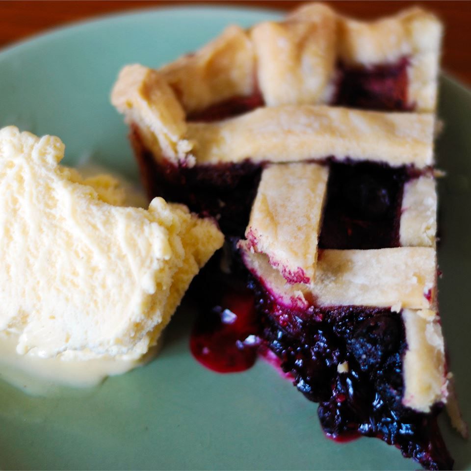 a slice of lattice-topped blueberry pie on a green plate with a scoop of vanilla ice cream