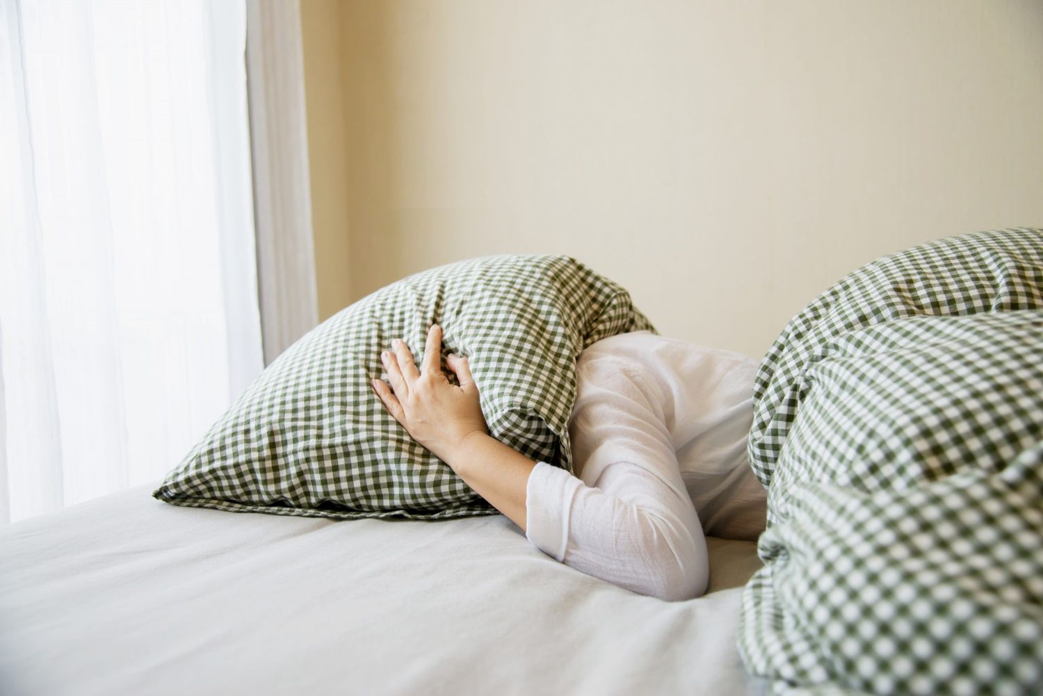 Young Woman Covering Pillow Lying On Bed Against Wall At Home