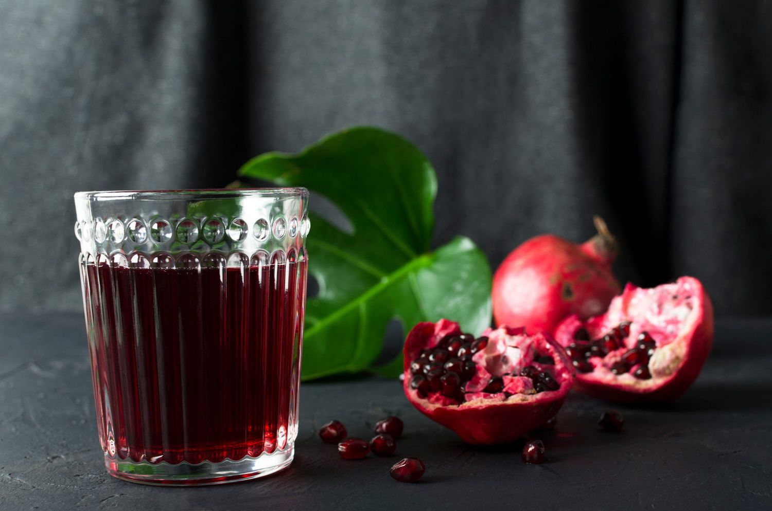 Stylish glass of delicious pomegranate juice, pomegranate seeds and tropical leaf on the dark table against grey background