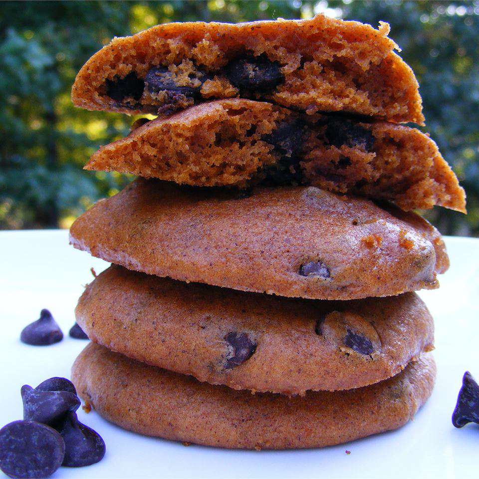 A stack of cake-like cookies, one broken in half, on a white plate scattered with chocolate chips