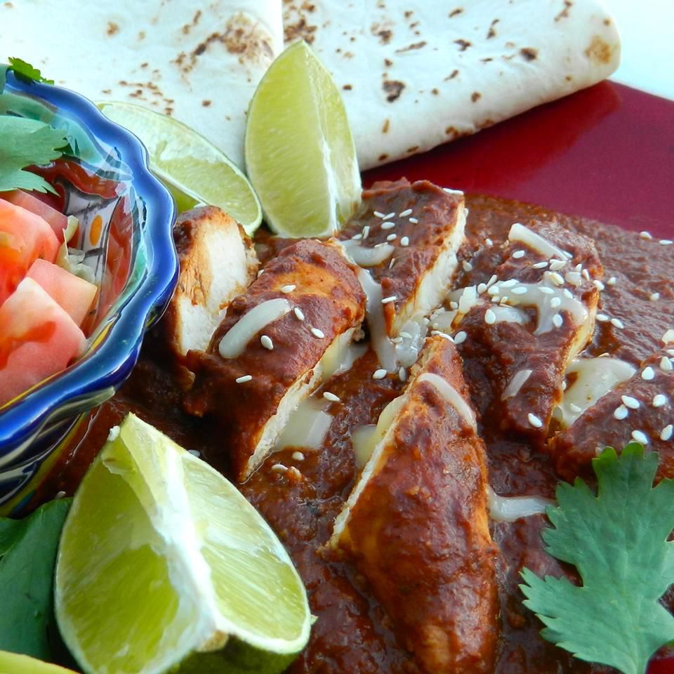 sliced chicken breast covered in a rich-looking brown mole sauce with lime wedges, pico de gallo, and flour tortillas on the side