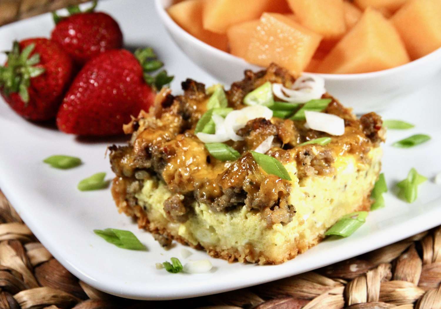 a square of sausage and egg casserole with a crescent roll base, topped with Cheddar and green onions, served on a white square plate with strawberries and cantaloupe behind it