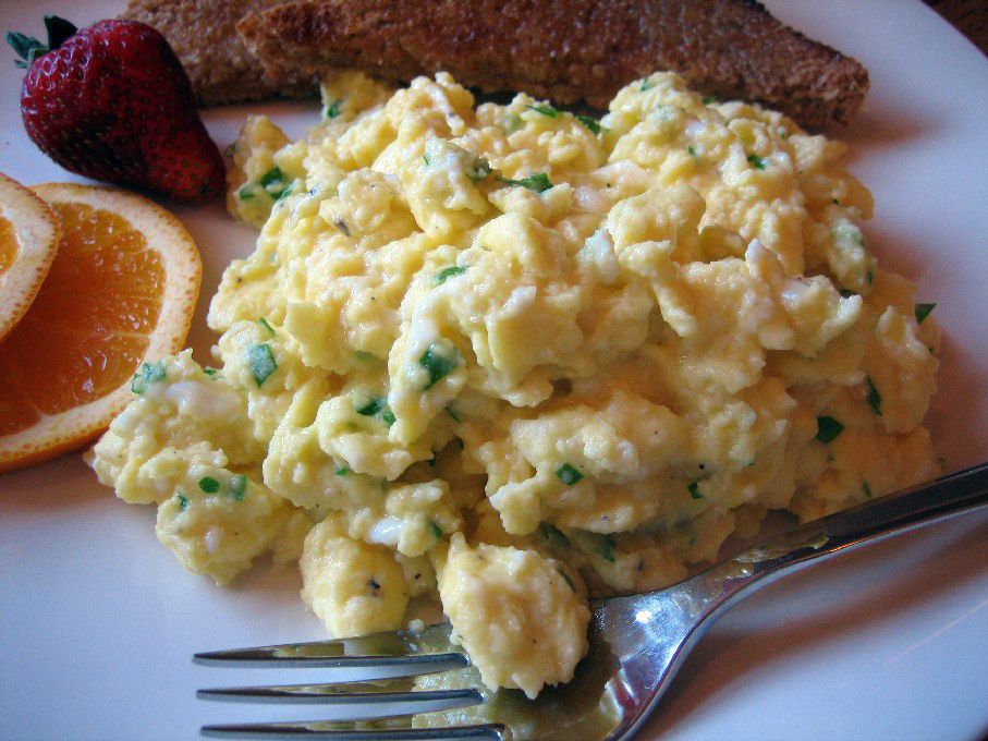 A serving of scrambled eggs on a white plate with toast and an orange garnish in the background