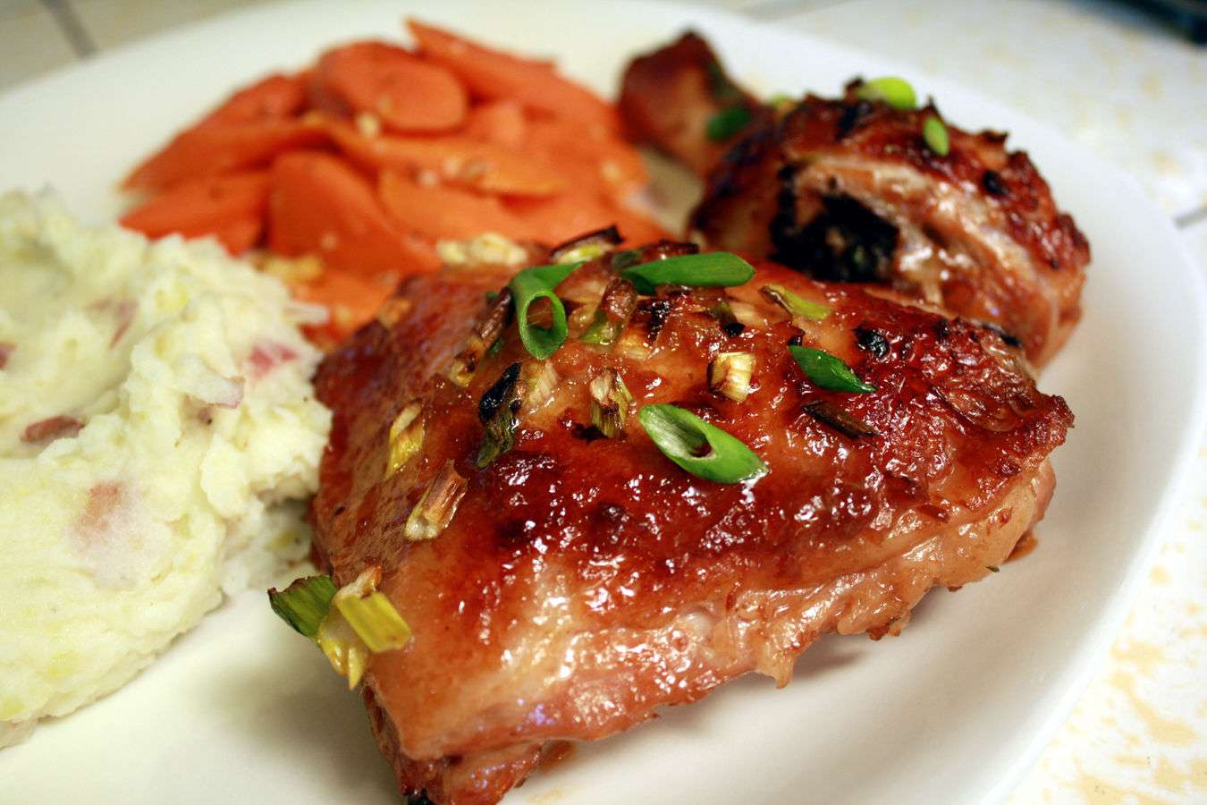 a dinner plate with a sticky-looking baked chicken piece topped with slivered green onions in the foreground, and mashed potatoes and glazed carrots in the background