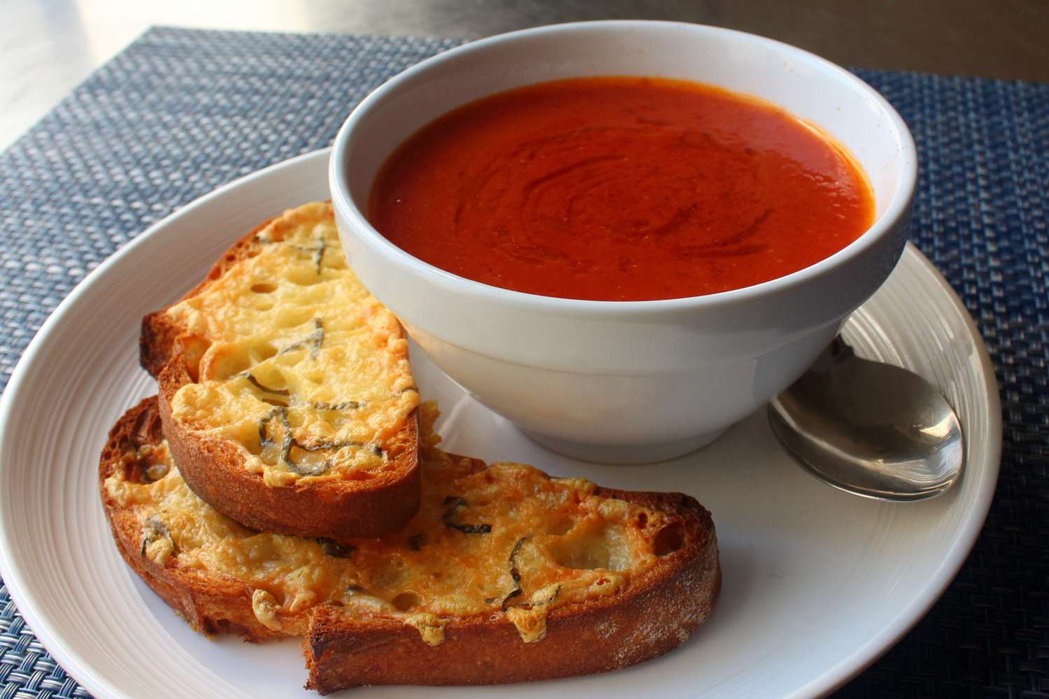 a white ceramic bowl of tomato soup on a matching plate with two cheese-topped baguette slices