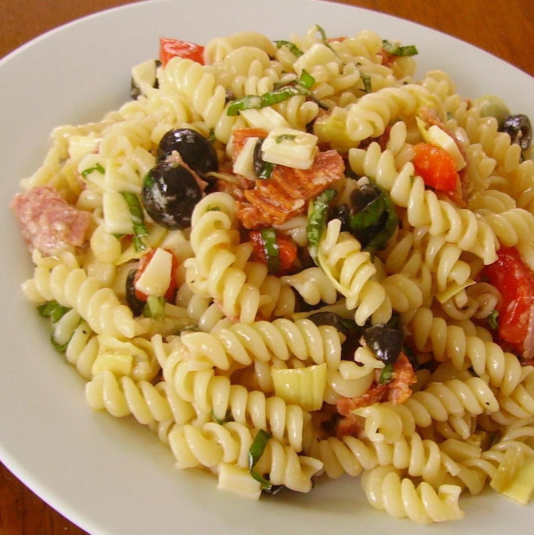 fusilli pasta on a white plate with black olives, salami, and roasted red peppers