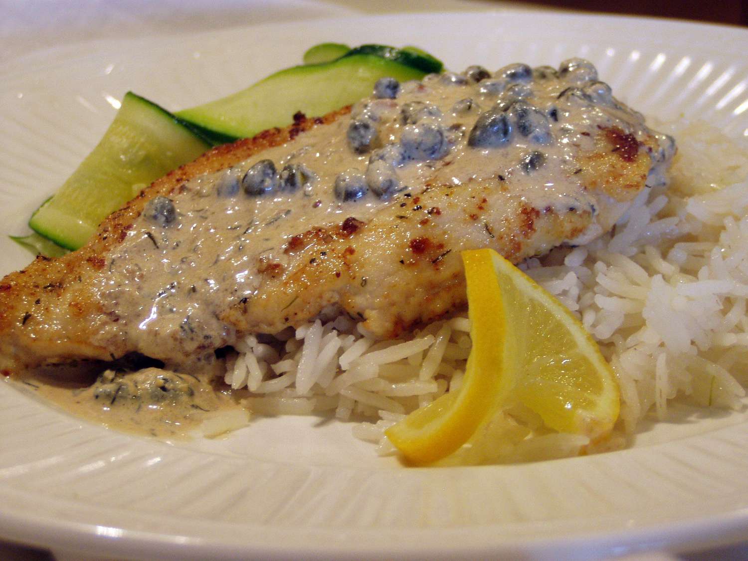 a chicken breast on a bed of white rice topped with cream sauce and garnished with a twisted lemon slice, with cooked zucchini slices in the background
