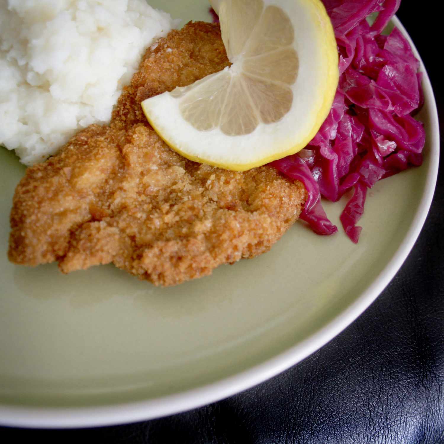 a dinner plate with a fried breaded veal cutlet garnished with a lemon slice, with red cabbage and mashed potatoes in the background