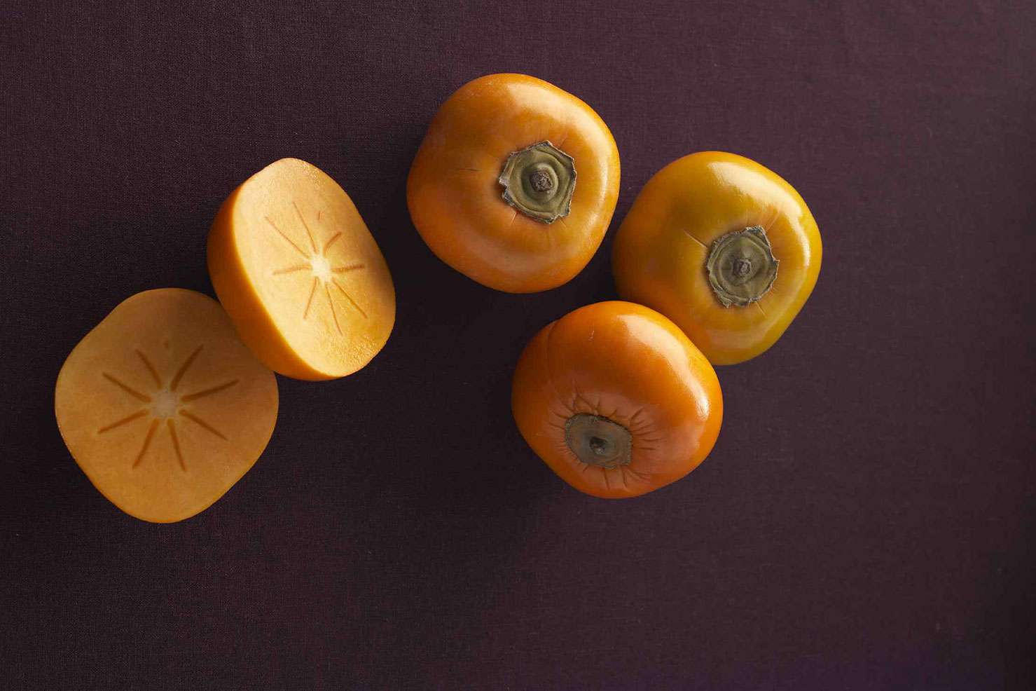 Three whole persimmons and one sliced one on brown/purple background
