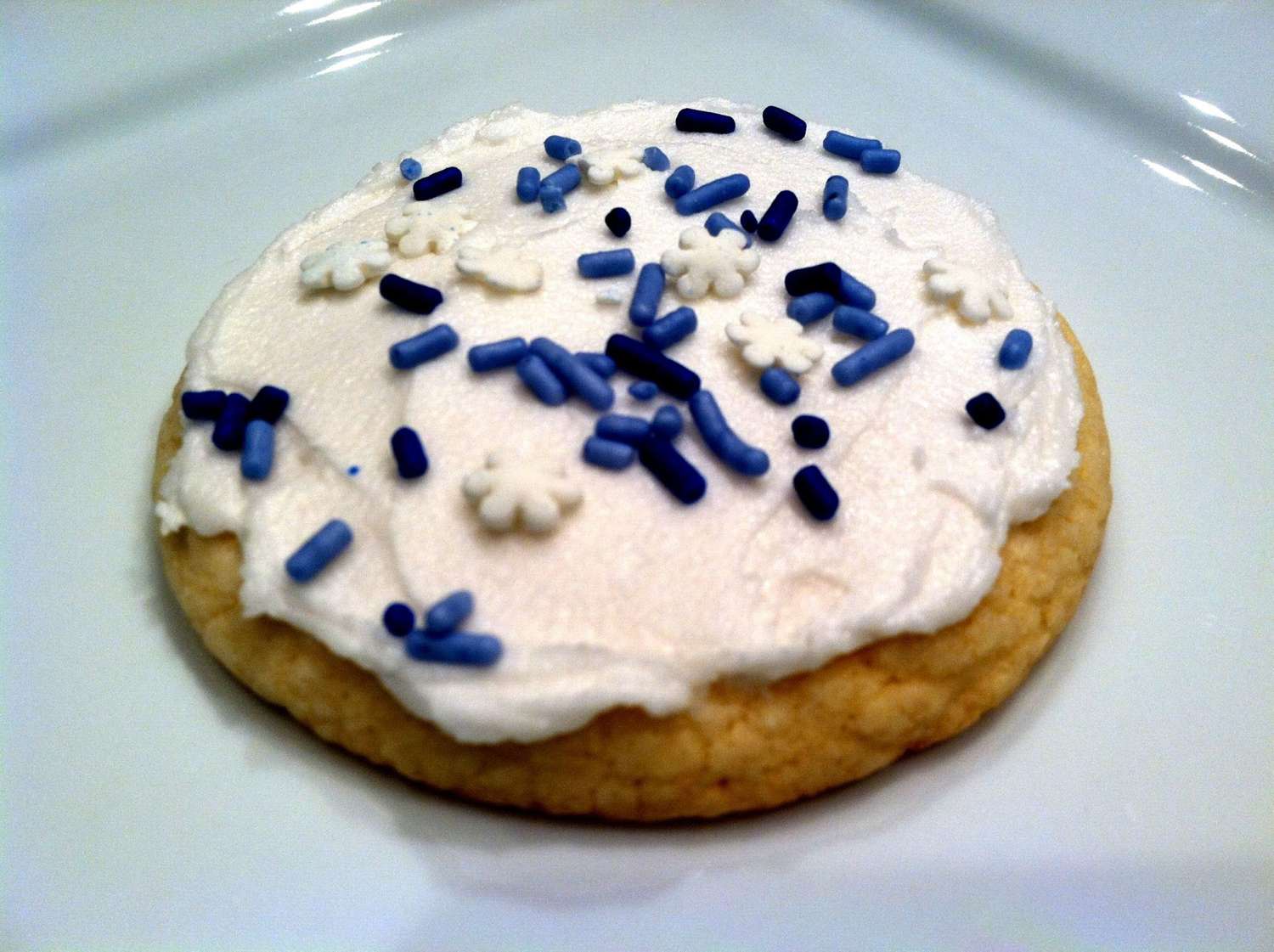 a round sugar cookie topped with creamy-looking white frosting and blue and white snowflake sprinkles