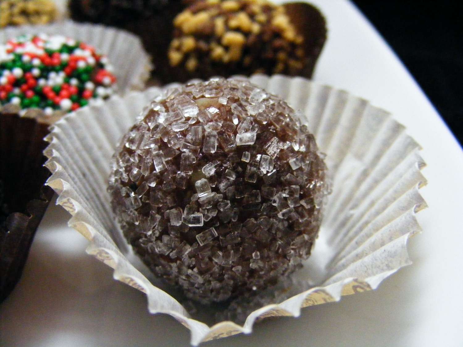 closeup of a rum ball rolled in sparkling sugar in a paper candy cup, with others rolled in different coatings in the background
