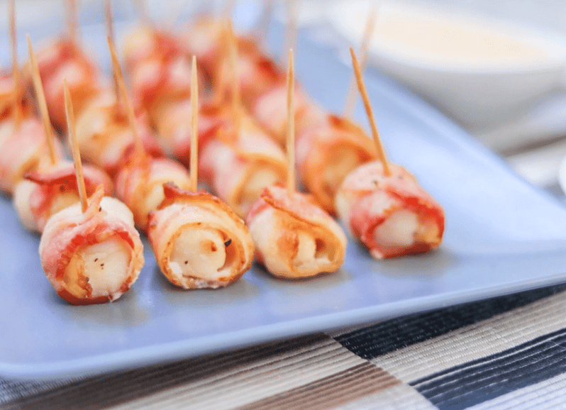 side view of rows of scallops wrapped in bacon secured by toothpicks on a square blue plate