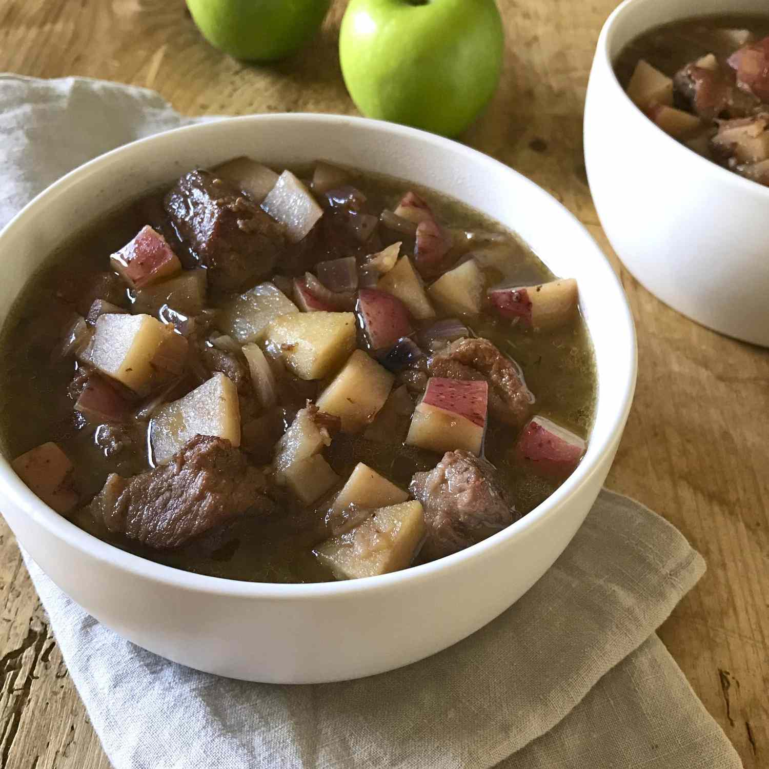Close-up on a bowl of pork stew with green apples in the background.