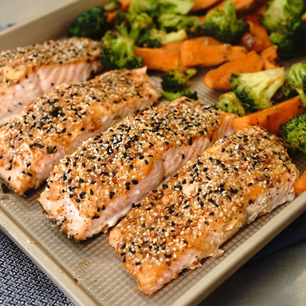 four salmon fillets topped with "everything bagel" seasoning on a sheet pan with broccoli florets and sweet potato wedges