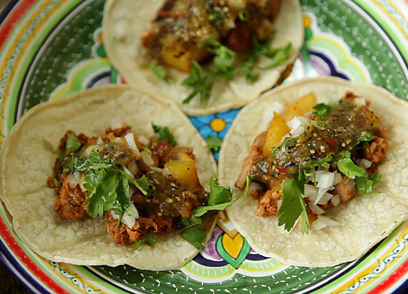 three tacos with pork, salsa verde, and cilantro on a painted ceramic plate