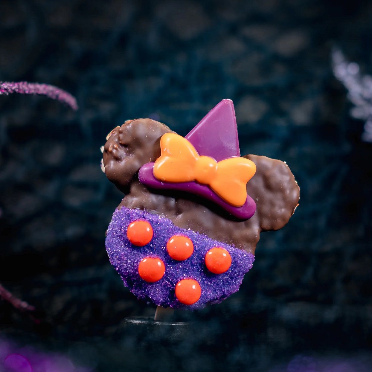 a chocolate covered crispy rice cereal treat cut out in the shape of a Minnie Mouse head wearing a witch's hat and decorated with purple and orange candies to create a Disney Halloween treat