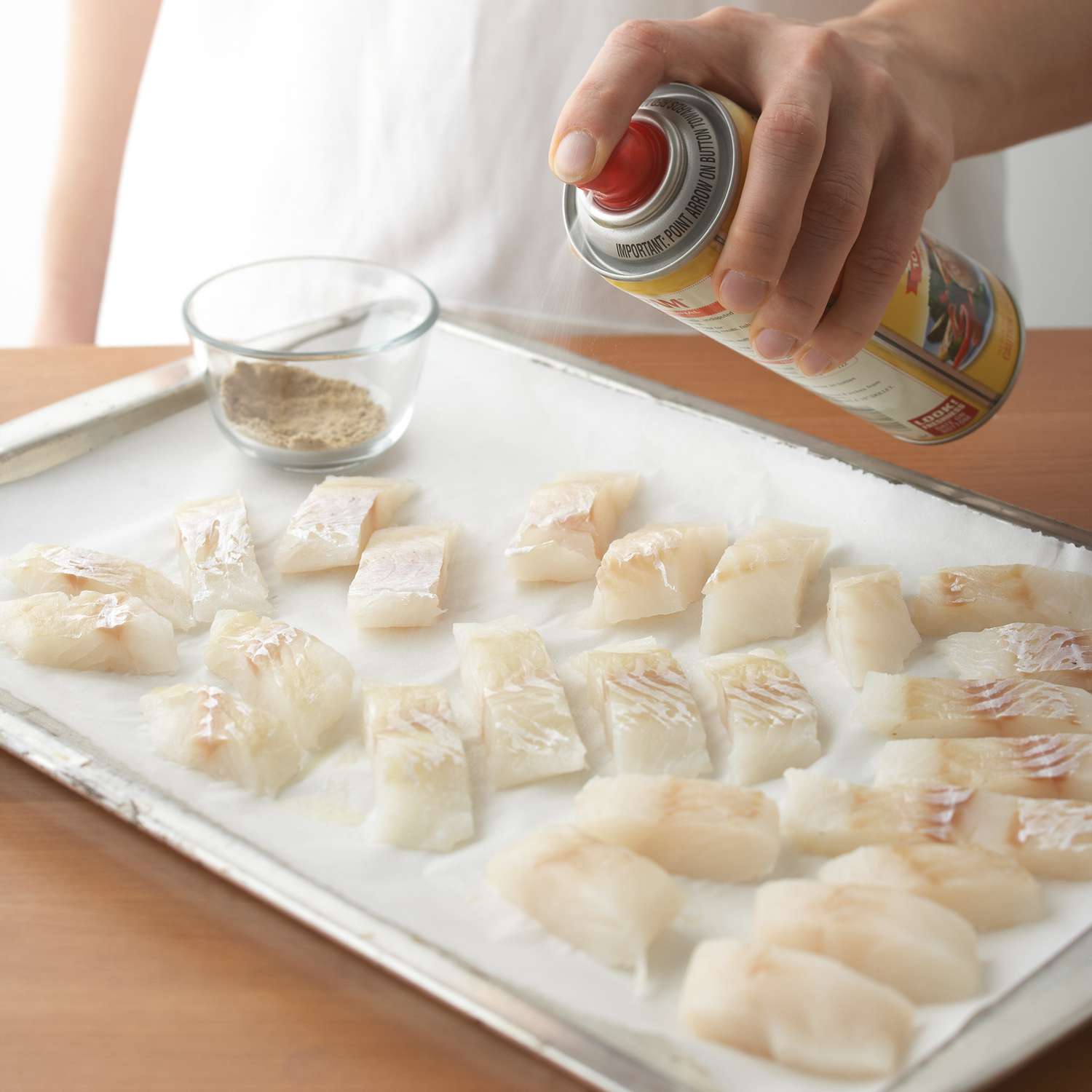 person spraying white filets with cooking spray on baking sheet