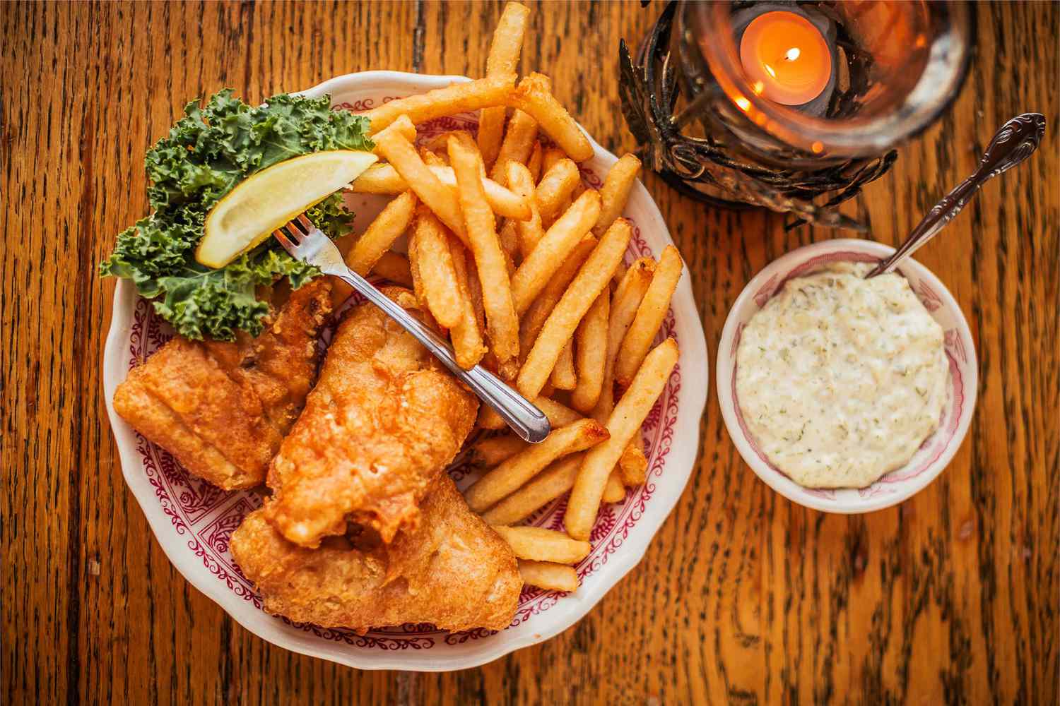 plate with lemon wedge, fork, fried battered fish, and fries next to bowl with tartar sauce and candle, all on wooden table