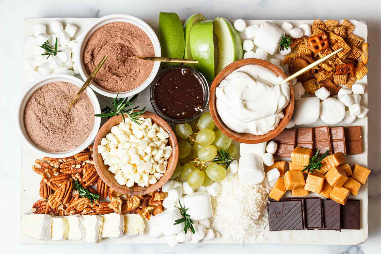 looking down at a hot chocolate charcuterie snack board with hot cocoa mixes, white and dark chocolate pieces, marshmallows, brie bites, nuts, snack mix, fruits, and whipped cream