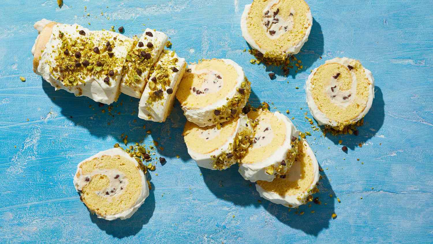 Slices of a cream-filled jelly roll cake topped with pistachios and chocolate chips on a blue surface