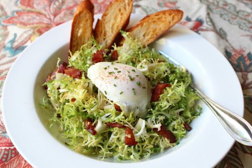 Salad of frisee lettuce, bacon, and a poached egg in a white bowl with three crostini