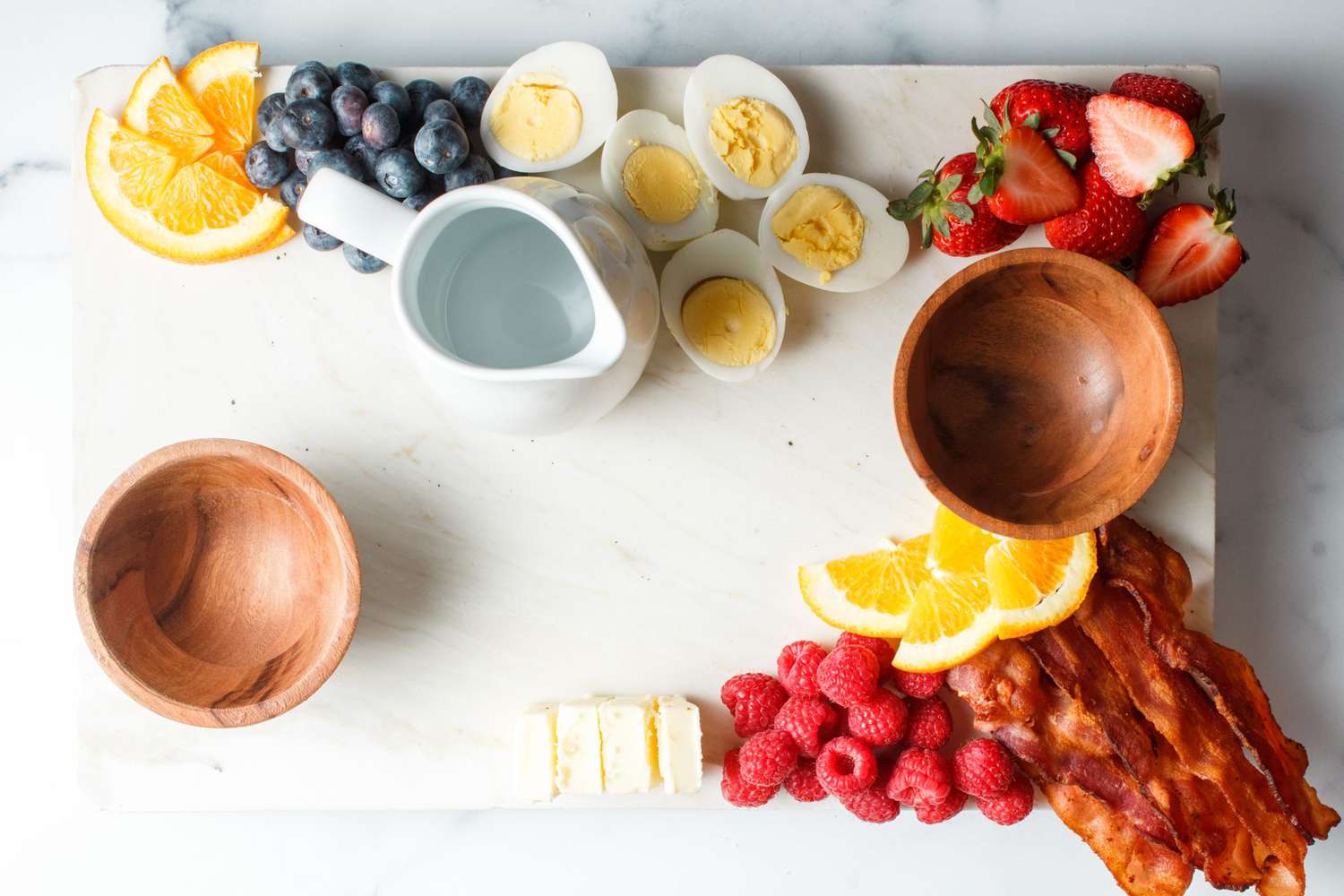 overhead view of a waffle charcuterie brunch board build in progress with sliced oranges, strawberries, raspberries, bacon, hard cooked eggs, small wooden bowls, and a syrup carafe.