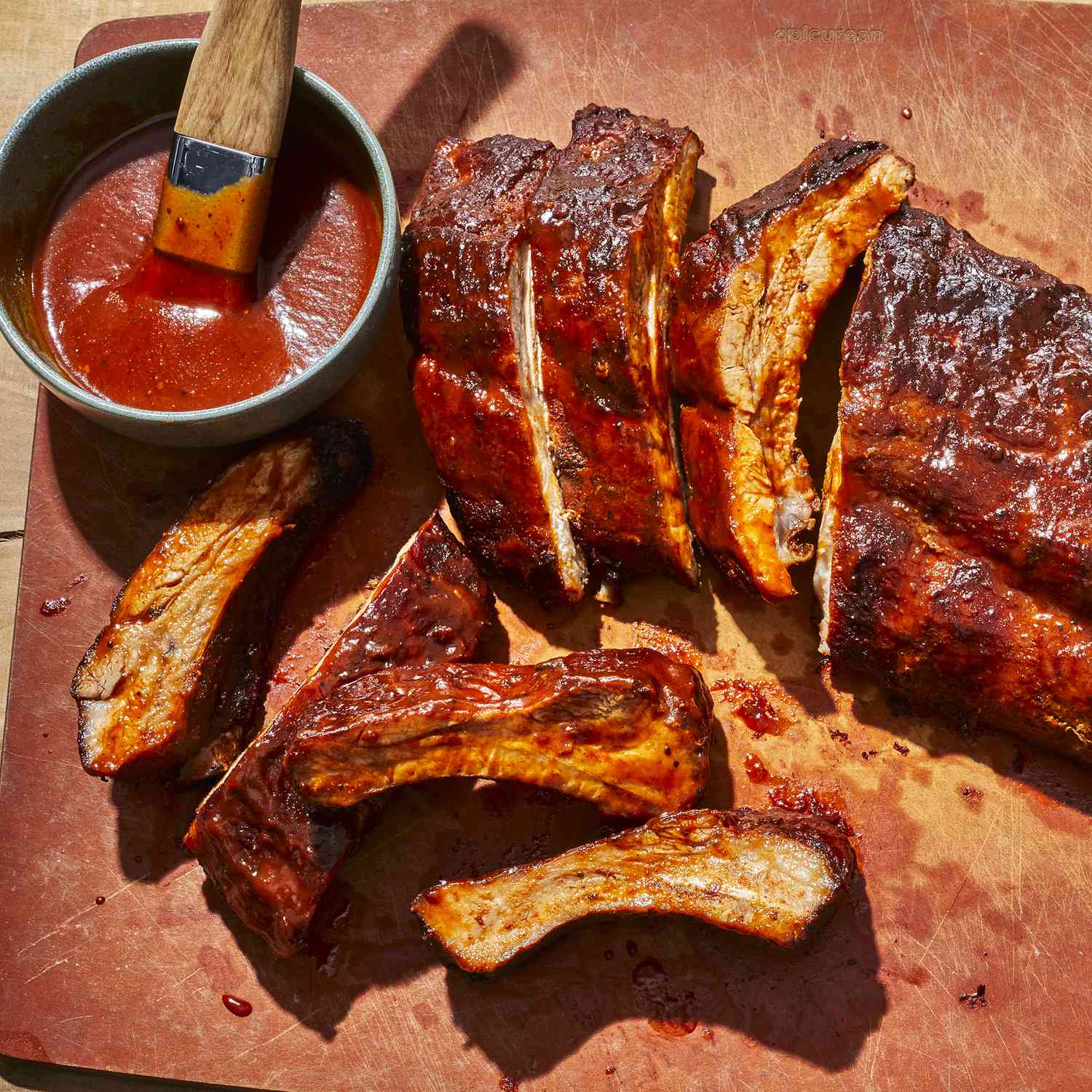 an overhead view of baked BBQ baby back ribs partially sliced on a cutting board with a ramekin of BBQ sauce nearby.