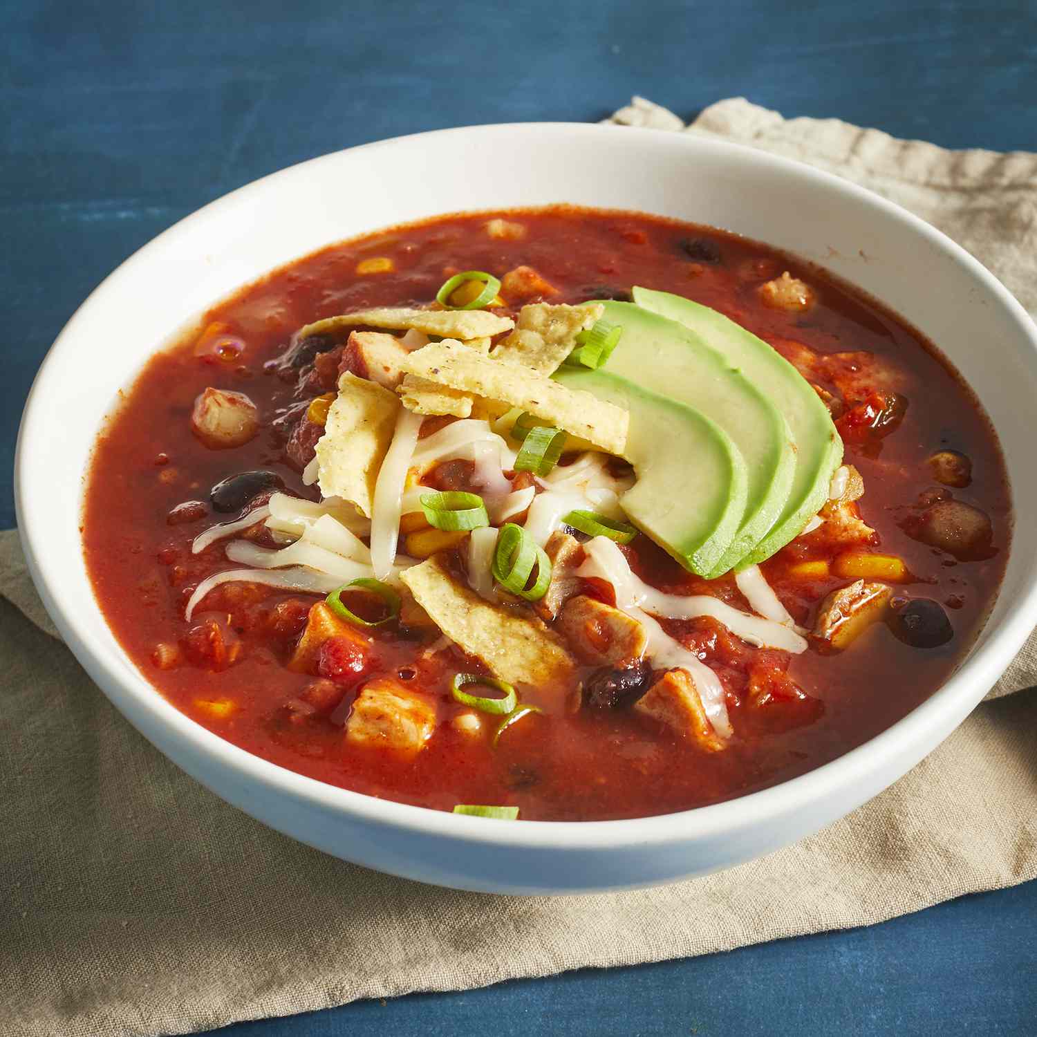 a low angle, close-up view of a single bowl of chicken tortilla soup topped with shredded cheese, crispy tortilla strips, and slices of avocado.