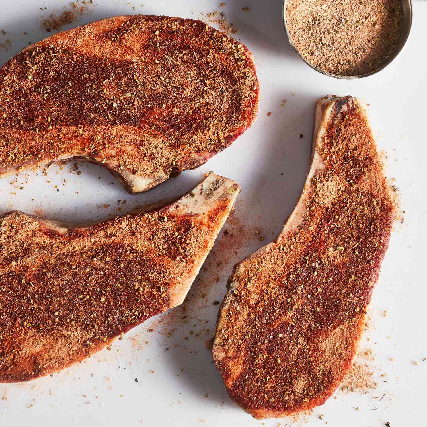 an overhead view of three raw steaks well seasoned with a dry rub mix of herbs and spices.